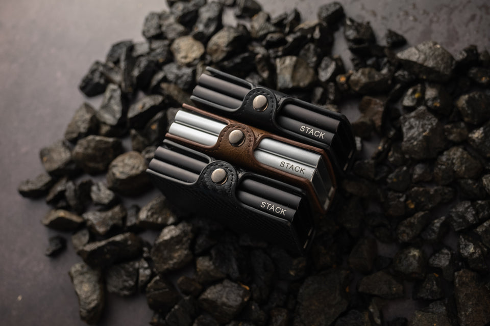 The Hs Kompact Stacks - Black, Dark Brown and Carbon Fibre wallets in a pile, placed on Black crushed rocks 